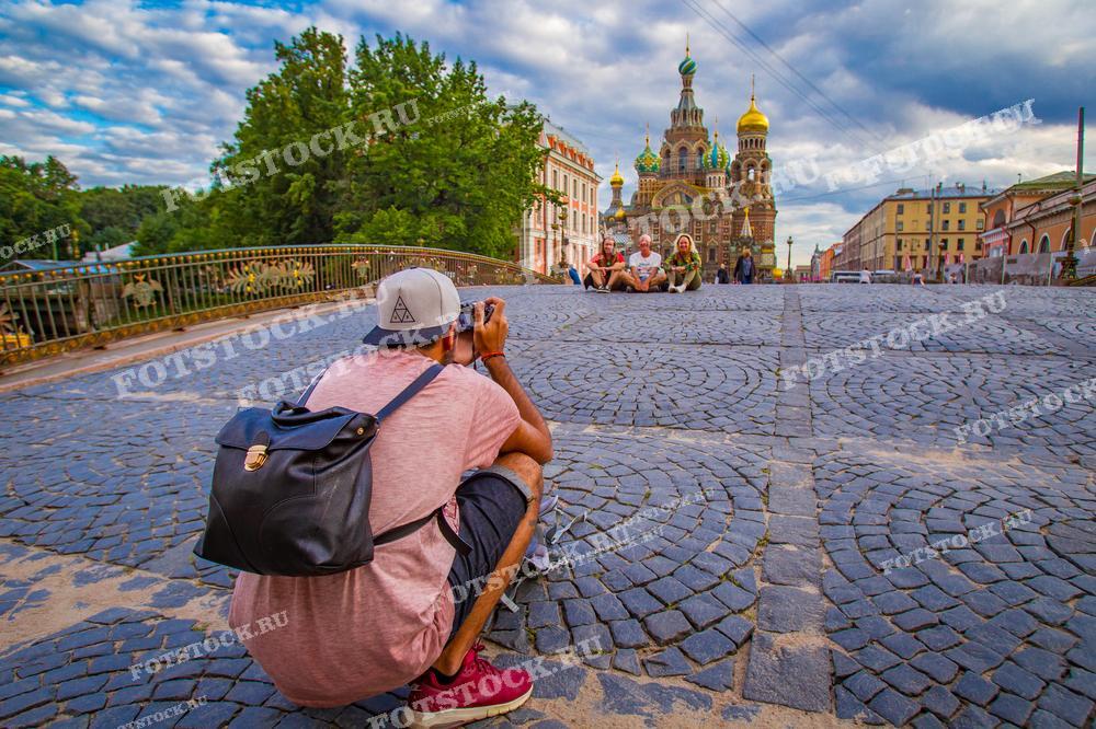 На фотографии изображён фотограф, сидящий на мостовой и делающий снимок. Он нацелен на Храм Спаса на Крови в Санкт-Петербурге, который виден вдали. На заднем плане на мосту расположились туристы, наслаждающиеся видами и отдыхающие. Мостовая и забор из кованого железа вносят в композицию аутентичный городской элемент. Небо украшено переменчивыми облаками, добавляющими яркости сцене. Такое фото может быть использовано в туристических брошюрах, статьях о путешествиях и как иллюстрация к блогам о городской фотографии.